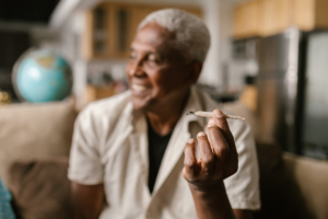 An older Black man sitting in a relaxed home setting, smiling while holding a cannabis joint. The image represents the role of cannabis in Black culture, holistic healing, and community wellness.